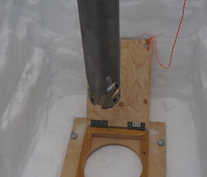 An ice core drill poised above ice. A wooden board with a circle cut in the centre marks the point where the drill will enter the ice.