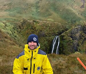 Man in yellow Antarctic Division jacket and blue beanie smiles at camera and give a thumbs up, in the background is Sawyer Falls which runs into a circular crater at base of tall grass covered hill