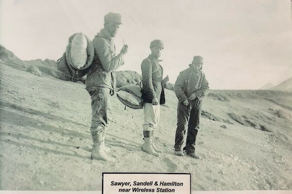 Black and white photo of three men standing on sloping beach, holding pipes and wearing historic back packs.