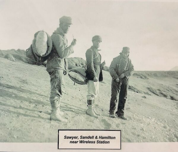 Black and white photo of three men standing on sloping beach, holding pipes and wearing historic back packs.