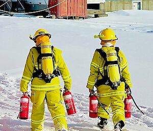 Two expeditioners in full fire fighting gear walk away from the camera angle on the snow to a building, both carrying two red fire extinguishers.