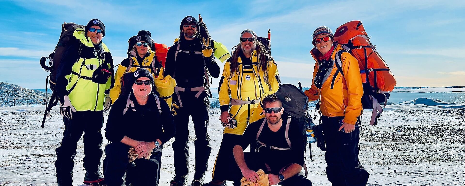 Seven expeditioners with their field packs and other equipment on the ice with water in the background