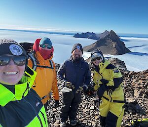 A group of people stand on a rocky hill on a blue sky day with lots of snow and ice and a few jagged hills in the background