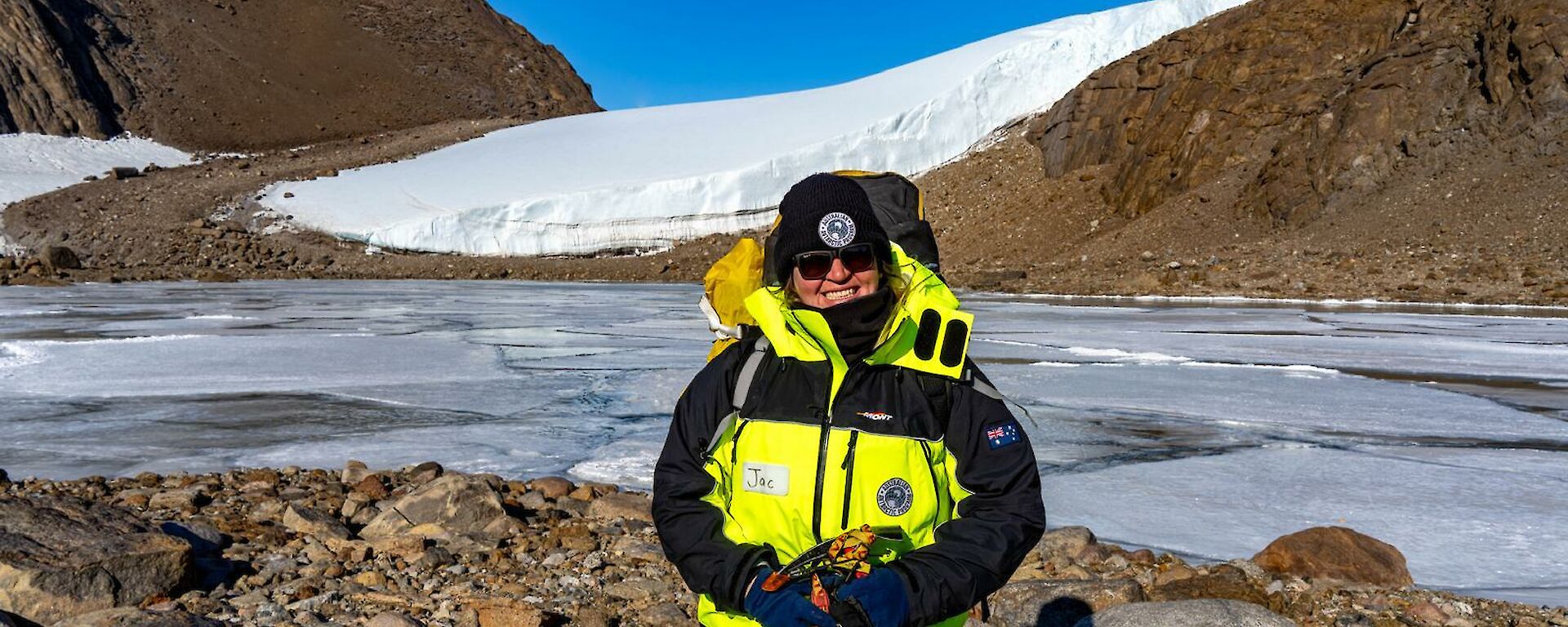 An expeditioner in yellow outdoor gear stands in front of a frozen lake with brown sheer hills in the background and blue sky