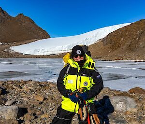 An expeditioner in yellow outdoor gear stands in front of a frozen lake with brown sheer hills in the background and blue sky