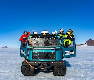 Three people leaning out of a metallic green hagglund, or snow tractor