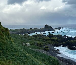 looking down a steep grass tussock slope to wooden hut in distance with two rocky promontories sticking out from coast line