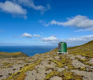 VHF communications repeater shelter, looks like a water tank with solar panels on the side, sits on windblown mountain top with steep drop off to the ocean in the distance, blue sky with small cumulous clouds above