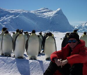 A man in red outdoor gear sits on the ice with emperor penguins standing behind him