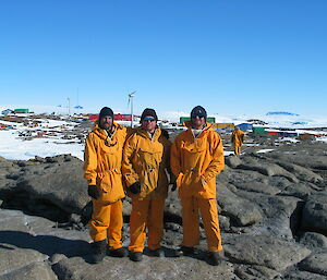 Three expeditioners stand in orange body suits dressed for the cold