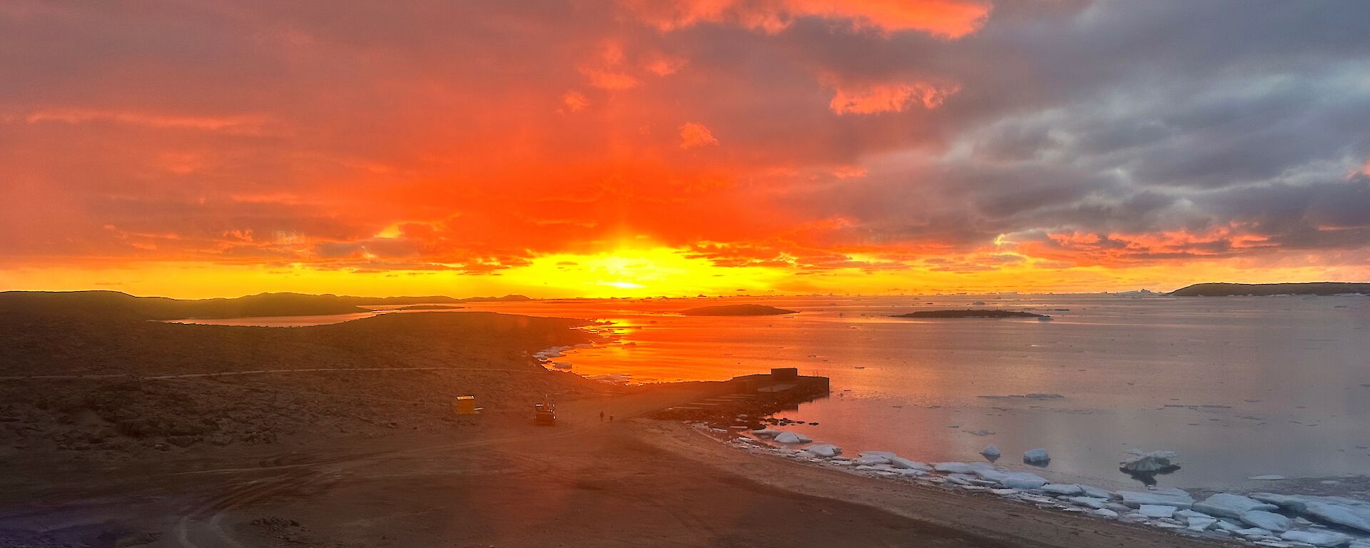 An orange sunset over a brown rocky landscape with water and ice in the background