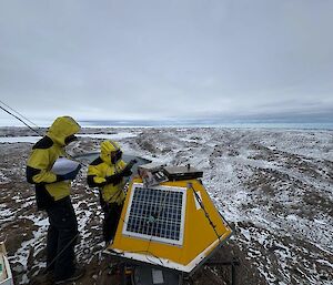 A person in yellow outdoor gear examines a yellow box with a solar panel on it, with icy hills in the background