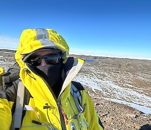 A person in yellow outdoor year wearing sunglasses, standing on a brown rocky hill with icy streaks over it