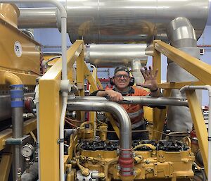 Man standing between two diesel generators smiles and raises one very dirty hand palm forward to camera