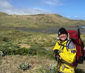 Man in foreground, smiling to camera, wears yellow jacket, black beanie and carries field pack. In the distance grass slope down to black sand bay and hills to horizen.
