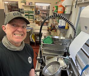 Man smiles to camera as he stands beside a huge pile of washing up in a kitchen sink