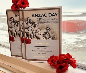 An Anzac Day program propped up against a window with red poppies around it