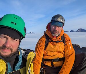 Two men in high vis and helmets sit on a rocky hill with ice behind them