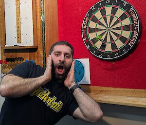 A man stands near a darts board looking shocked