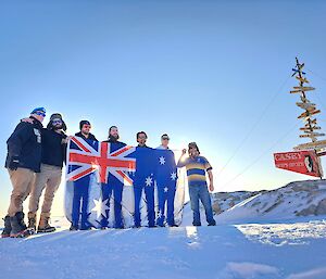 Expeditioners stand in front of the Casey sign holding an Australian flag.