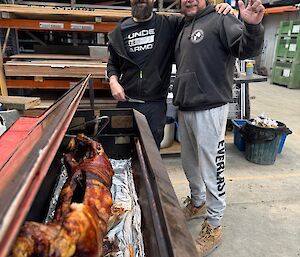 Two men in a warehouse cooking a pig on a spit