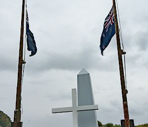 Australian flags at half mast in front of a make-shift white cenotaph and cross, on green ground