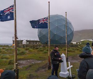 People gather under two Australian flags with a white cross in the ground and round satellite dome behind them