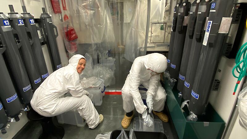 Two people in white hazmat suits in a lab with tall grey cylinders