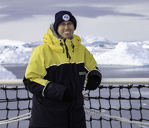 A man in yellow Antarctic wear stands on a ship's deck with icebergs behind him