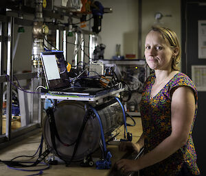 A blonde woman in short sleeves, stands in front of complicated looking equipment and a computer screen
