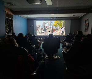 A view from the back of the movie theatre at Casey Station with a movie on the big screen and the expeditioners watching in darkness