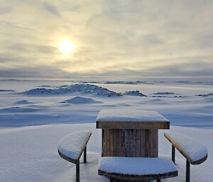 a picnic table covered in snow with the sun setting in the background