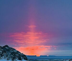 An orange sunset against a purple blue sky with a snowcapped rocky outcrop to the left and icebergs in the far distance