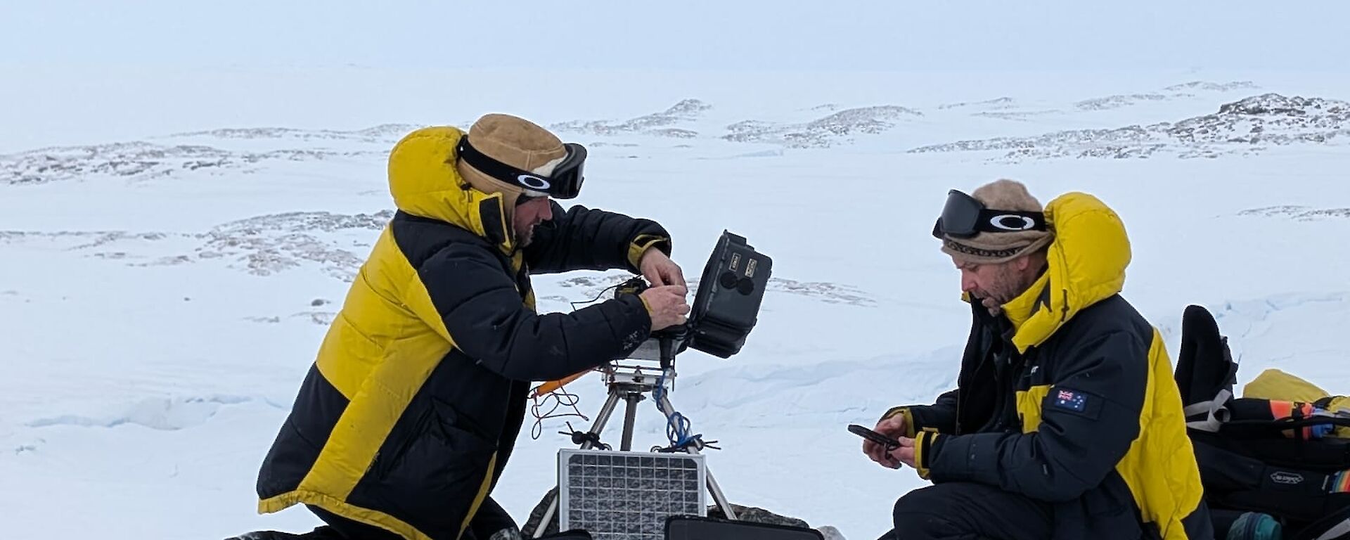 Two expeditioners servicing cameras and downloading data whilst  squatting down on the ice