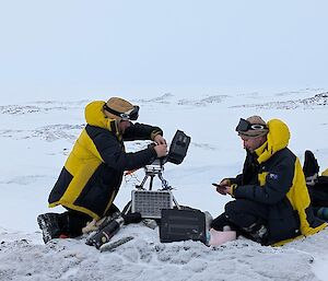 Two expeditioners servicing cameras and downloading data whilst  squatting down on the ice