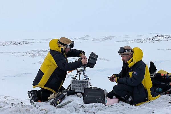 Two expeditioners servicing cameras and downloading data whilst squatting down on the ice