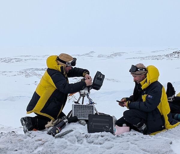 Two expeditioners servicing cameras and downloading data whilst squatting down on the ice