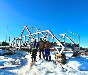Three people stand in the snow in front of a metal  bridge
