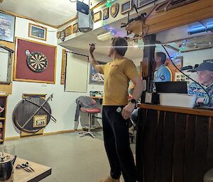 A man in a yellow tshirt lines up to throw a dart