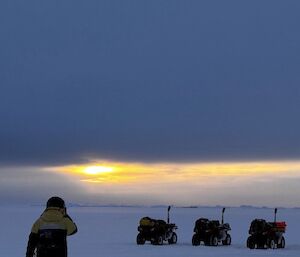 man walking on sea ice with three quad bikes in the background