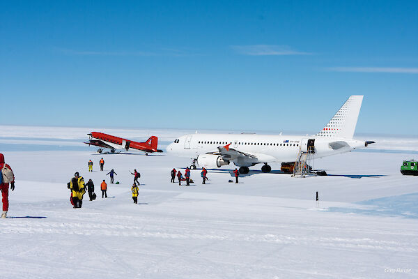 A big white plane on an ice runway with a smaller red plane to the left and lots of people in yellow walking away from them