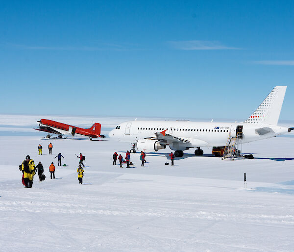 A big white plane on an ice runway with a smaller red plane to the left and lots of people in yellow walking away from them