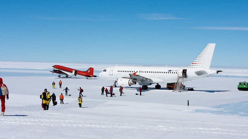 A big white plane on an ice runway with a smaller red plane to the left and lots of people in yellow walking away from them
