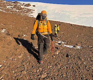 Two expeditioners walk across a rocky slope with ice and blue sky in the backgroud