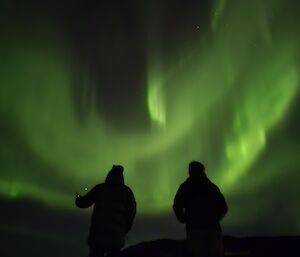 Two dark figures in front of a shimmering green aurora in the night sky