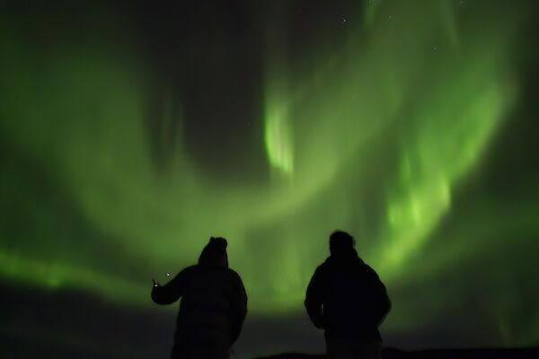 Two dark figures in front of a shimmering green aurora in the night sky