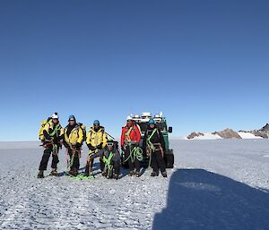 A group of people in yellow outdoor gar with a Hagg and search and rescue equipment on a white snowy plain with rocky hills in the background