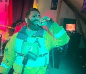 A man singing into a microphone wearing yellow outdoor gear