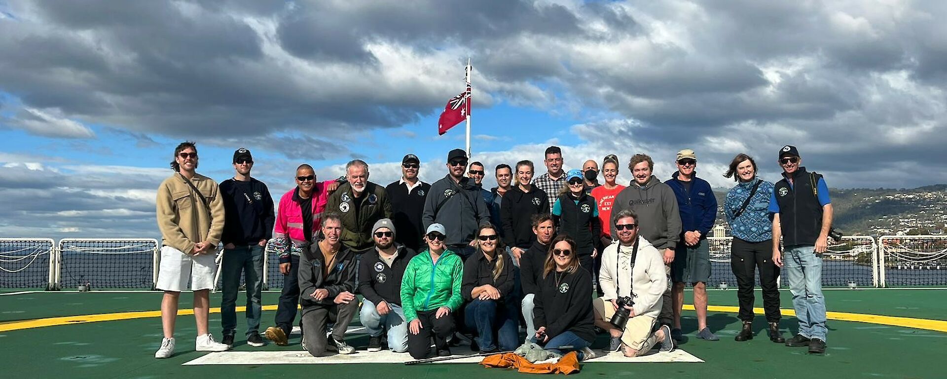 A group of smiling people standing in a group on a green helicopter deck on the back of a ship.