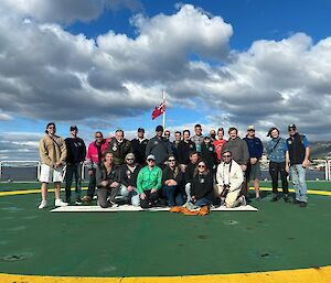 A group of smiling people standing in a group on a green helicopter deck on the back of a ship.